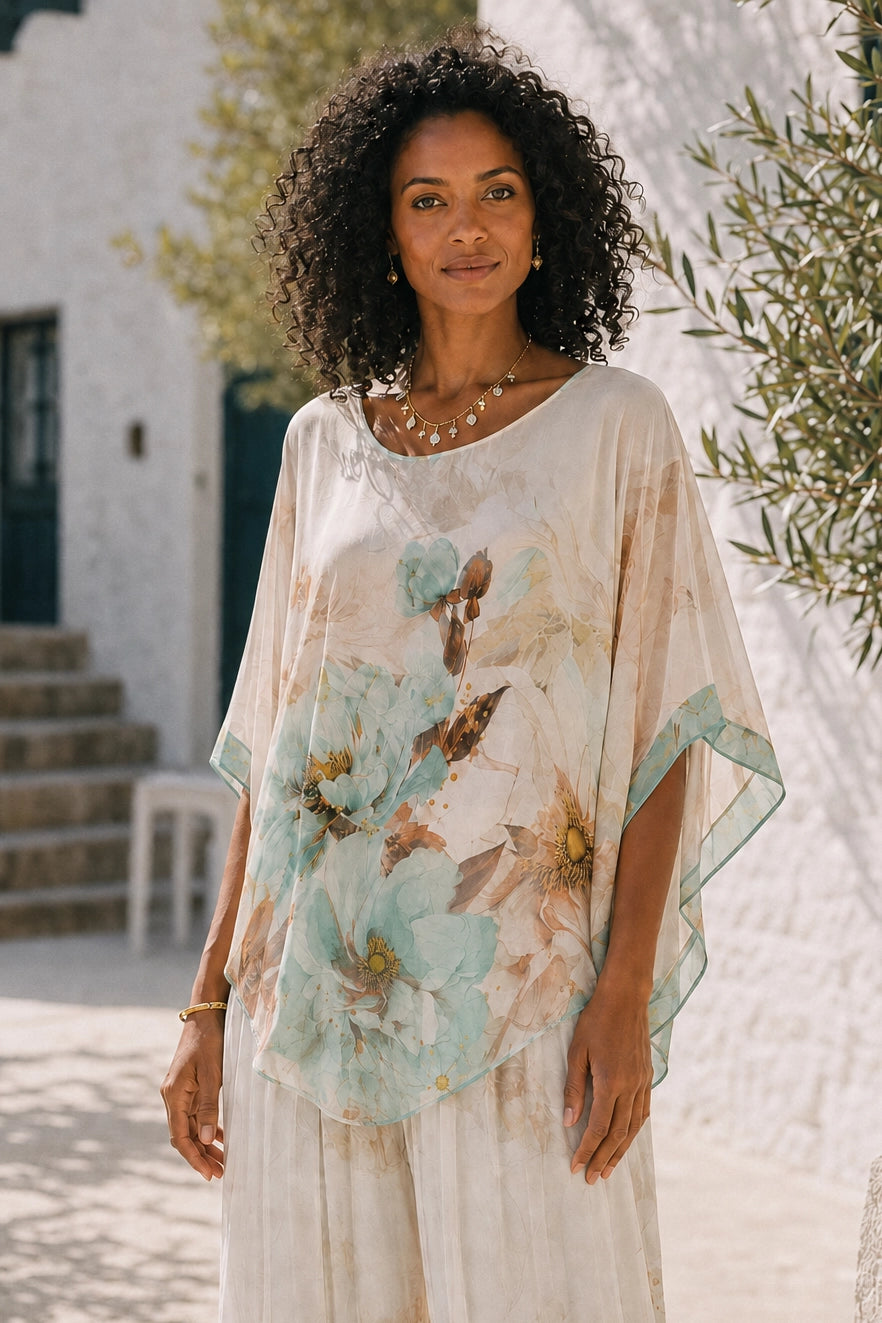 A woman with curly black hair wearing a flowing, semi-sheer cream kaftan dress with a large light blue and beige floral print, standing outdoors in front of a white stucco building.