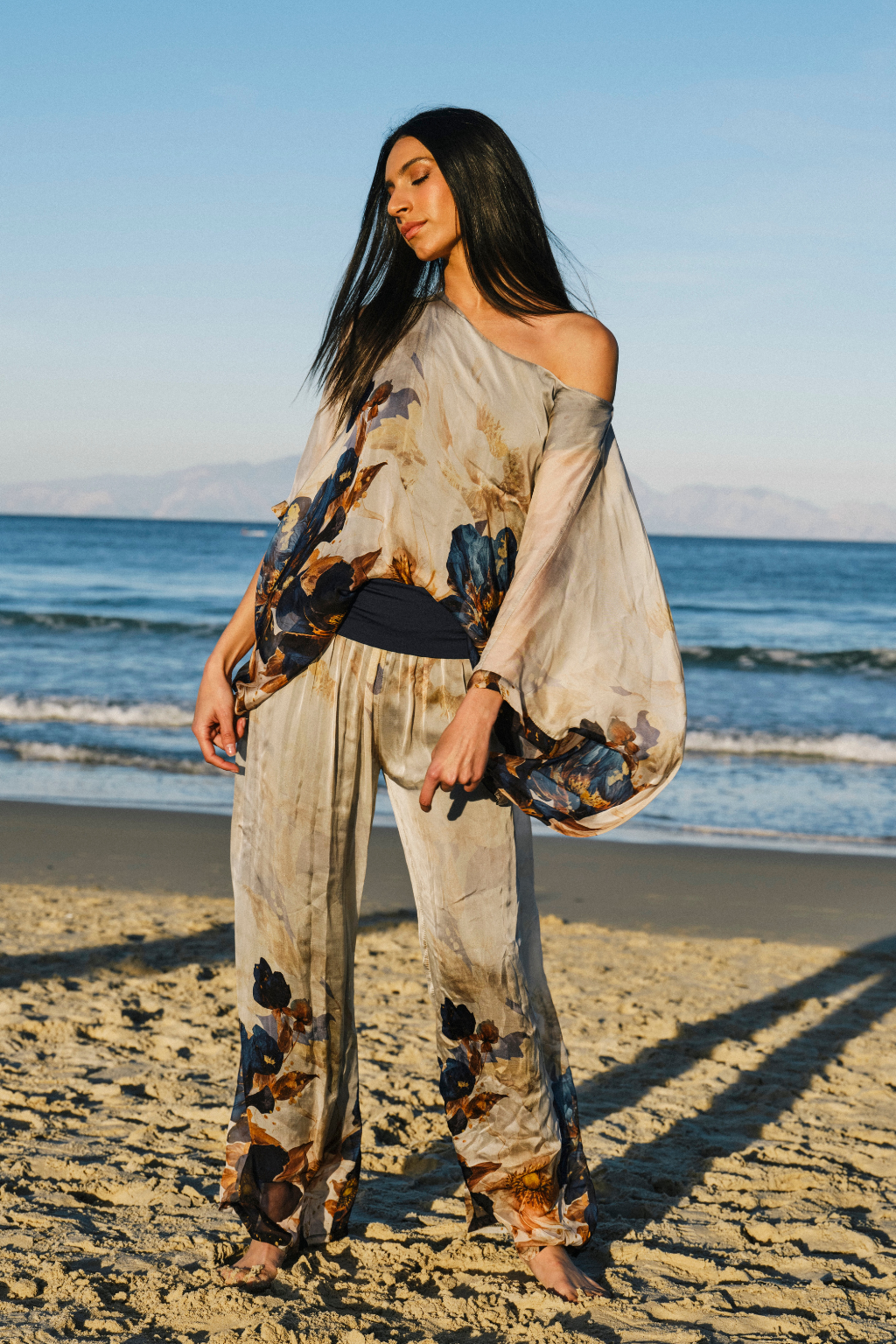 A woman stands on a sandy beach wearing a loose, one-shoulder beige jumpsuit with a dark blue and brown abstract floral print. The ocean and blue sky are visible in the background.