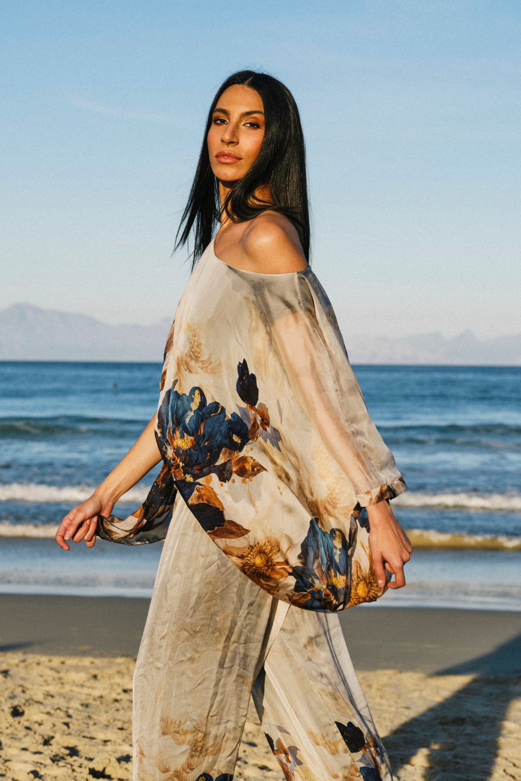 A woman stands on a sandy beach wearing a loose, off-the-shoulder maxi cover-up with a blue, brown, and beige floral print. The background features the ocean, distant mountains, and a clear blue sky.