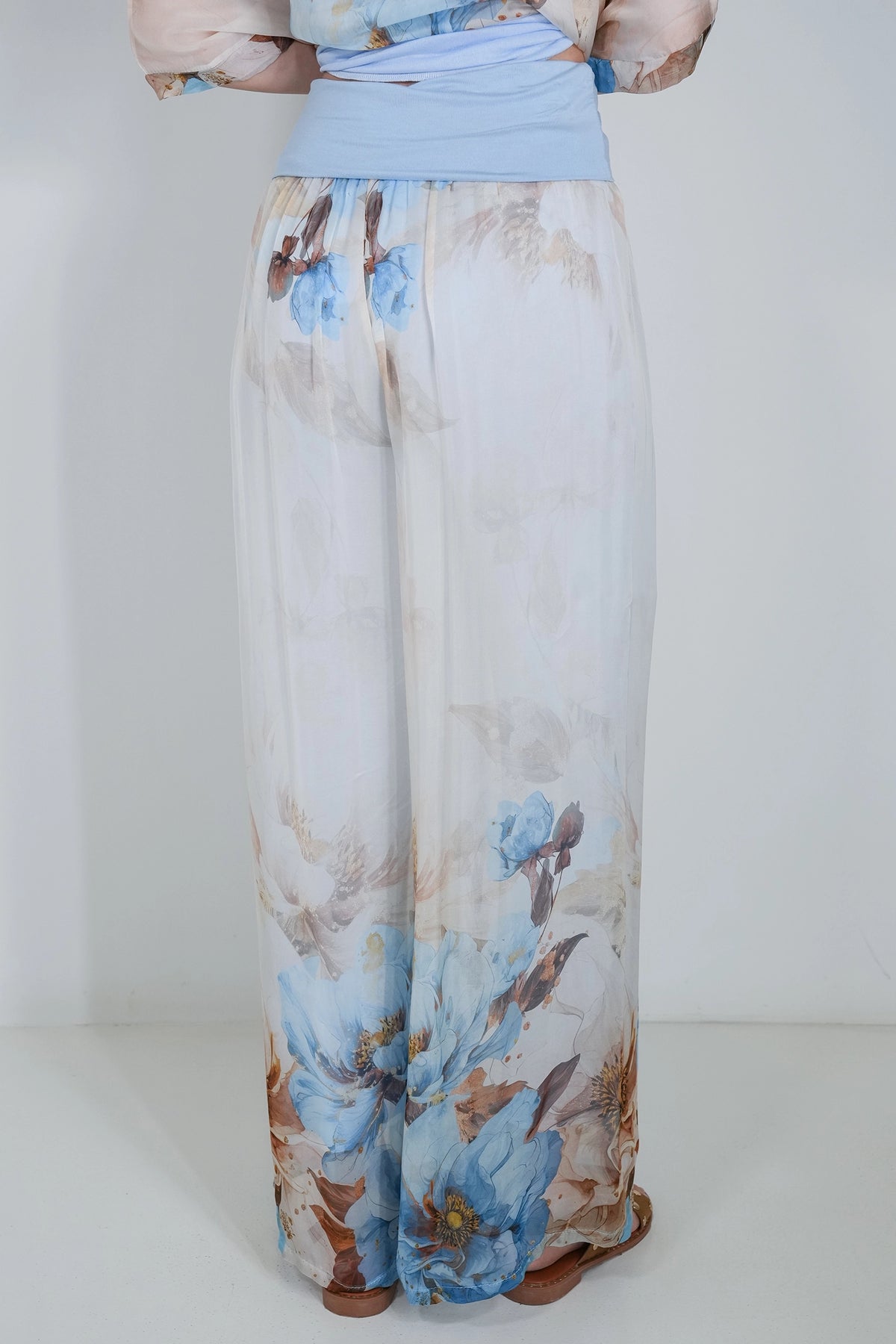 A person shown from the back wearing wide-leg pants with a white base and a blue and brown floral print, paired with a light blue waist wrap.