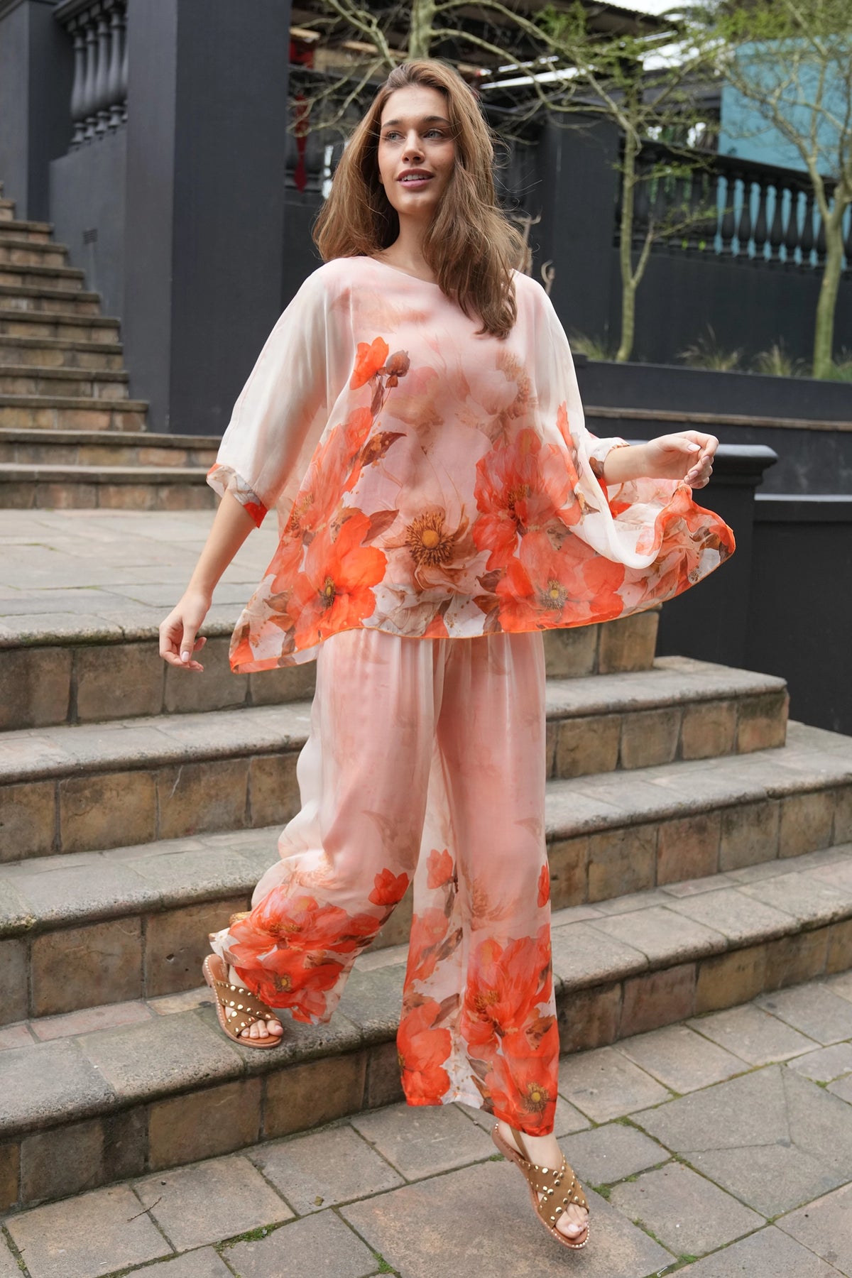 A woman wearing a sheer peach and orange floral jumpsuit with wide-leg pants and gold sandals, walking down outdoor stone steps.
