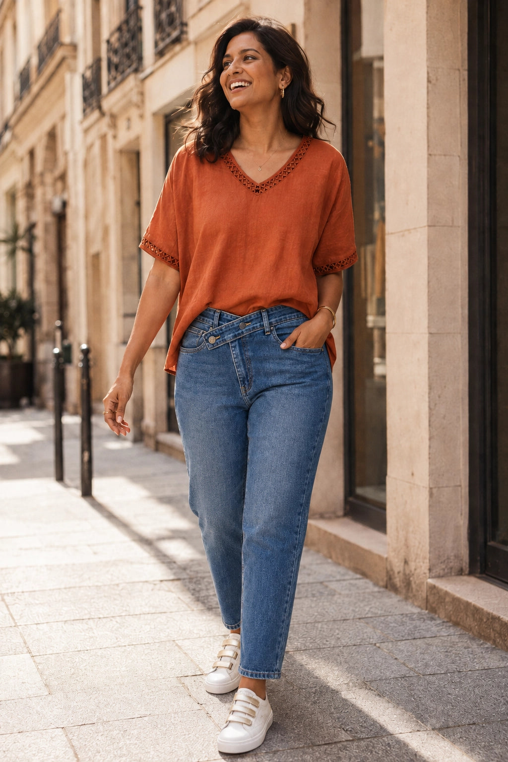 A woman walking on a sunny city sidewalk wearing a rust-orange short-sleeve top with lace trim, blue jeans, and white sneakers.