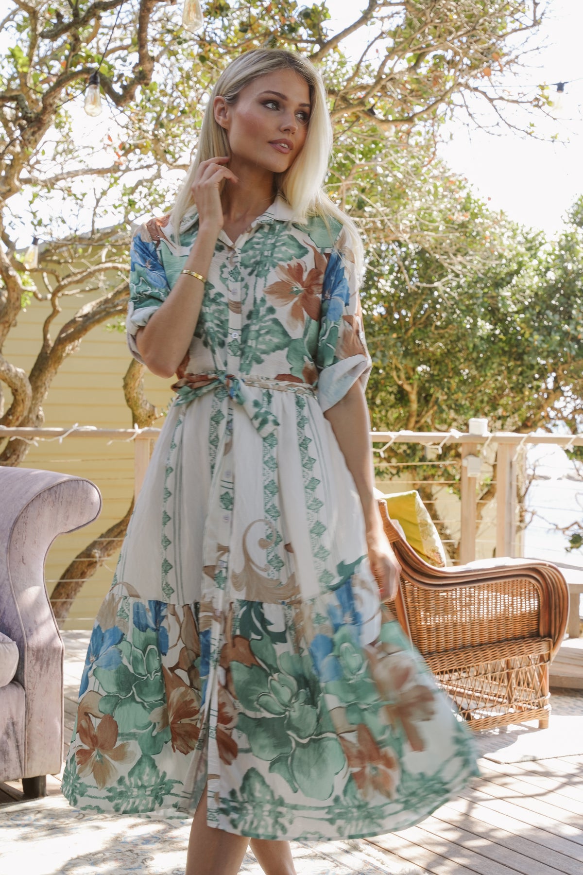 Woman wearing a floral dress standing outdoors with trees and furniture in the background