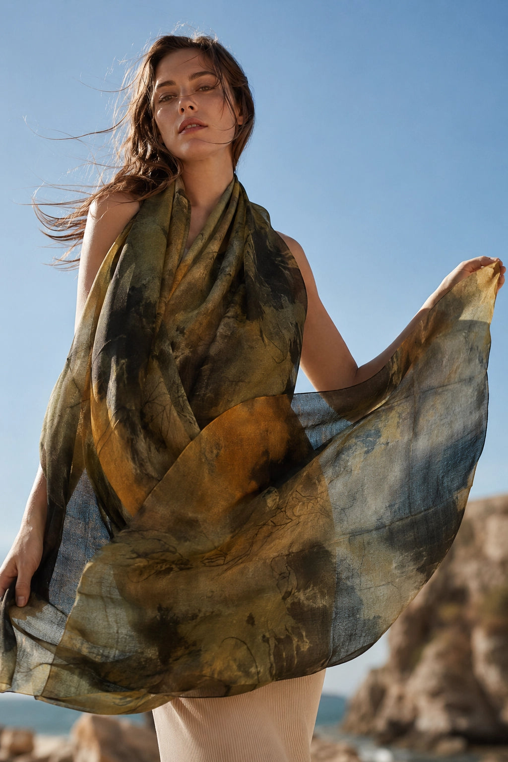 A woman outdoors by the sea wearing and holding a lightweight draped scarf with an abstract watercolor-like print in olive green, mustard yellow, brown, and blue-gray tones against a blue sky.