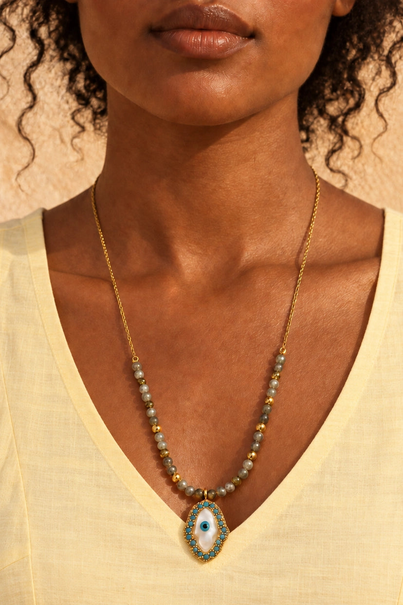 A close-up of a model wearing a gold chain necklace with light blue-gray beads and an evil eye pendant.