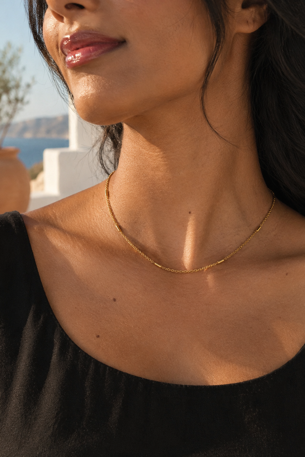 A close-up lifestyle shot of a woman wearing a delicate gold chain necklace, with a sunny outdoor background showing a glimpse of the sea and white buildings.