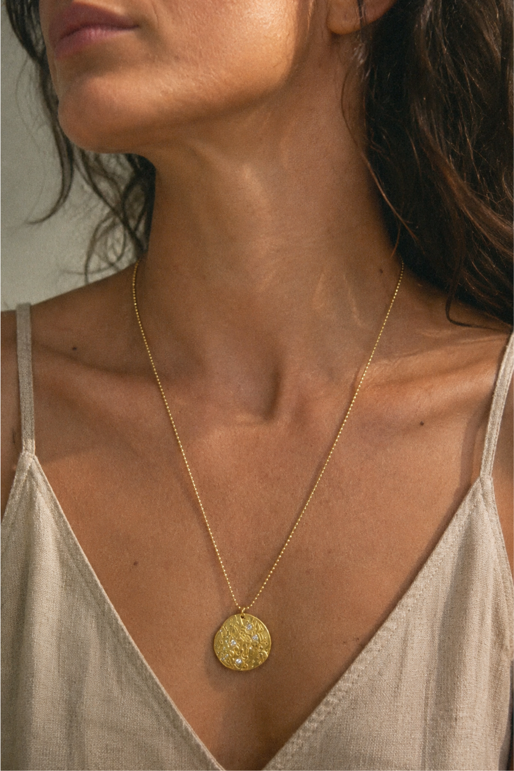 A close-up of a woman wearing a delicate gold chain necklace with a textured circular pendant, styled with a beige spaghetti strap top.