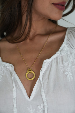 Close-up of a woman wearing a gold necklace with a circular pendant against a white blouse.