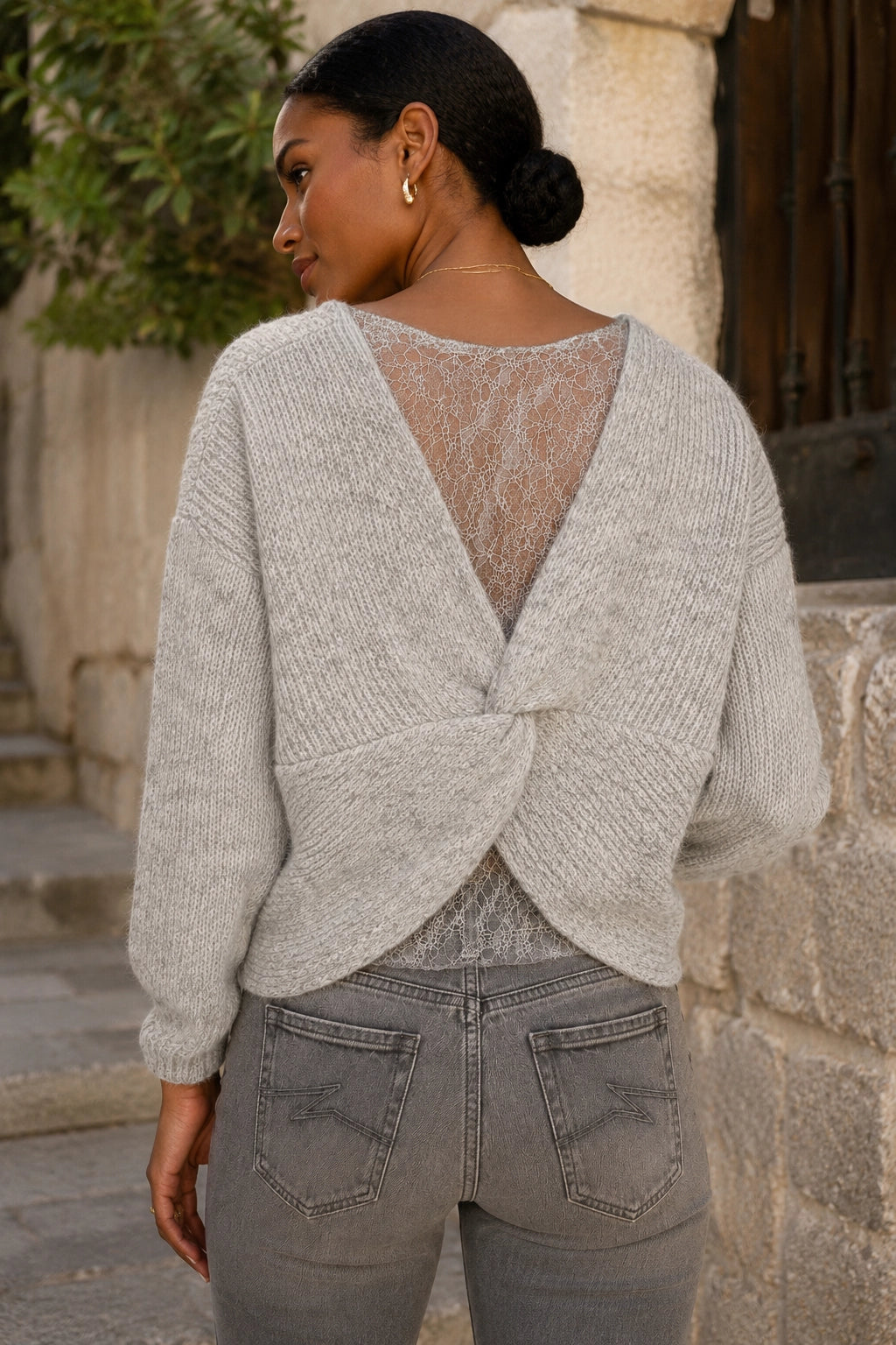 A woman wearing a light gray knit wrap cardigan with a twisted back detail and a lace insert, paired with gray jeans, photographed outdoors against a stone wall.