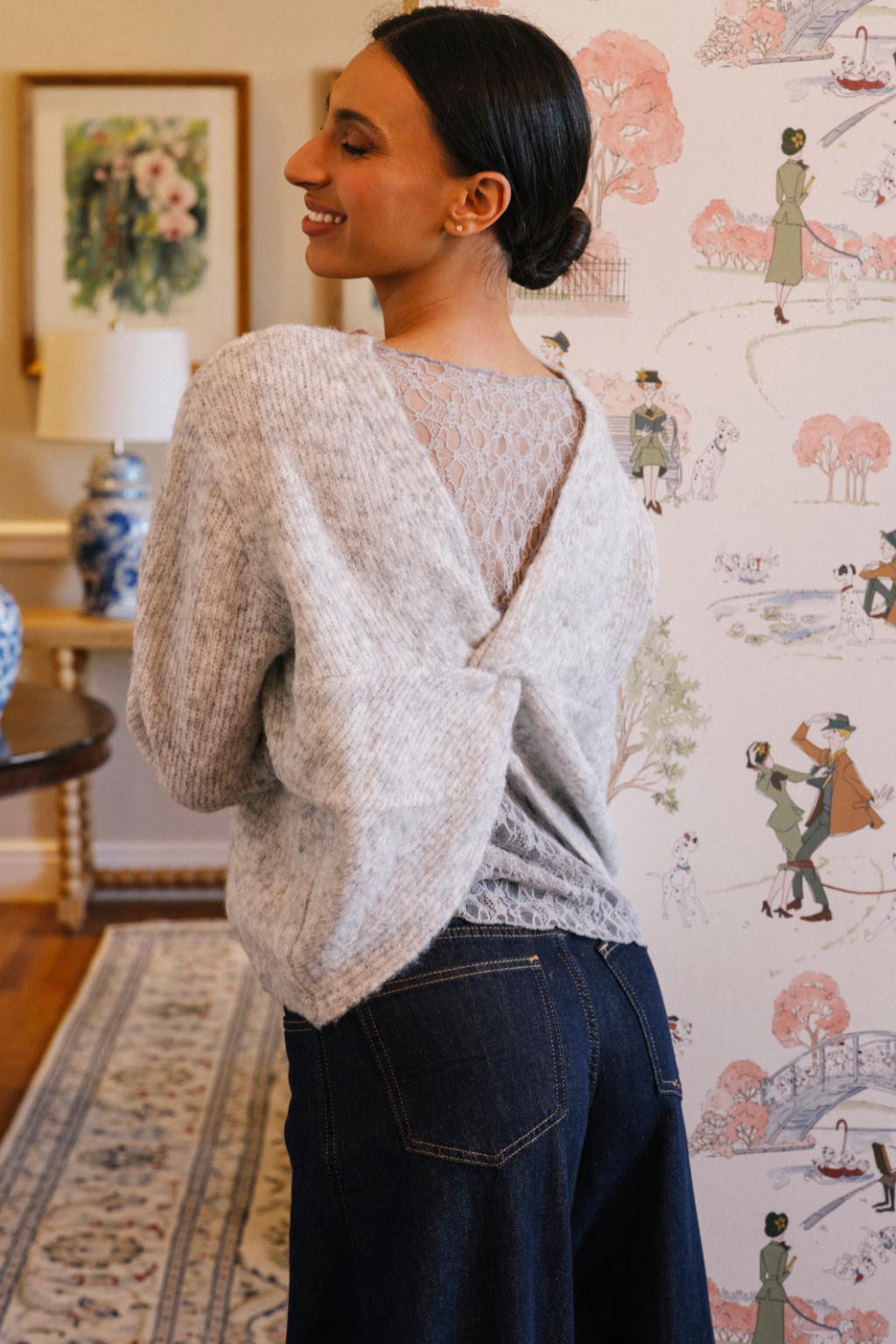 A woman wearing a light gray knit wrap sweater with a lace-trimmed open back and dark blue jeans, standing in a decorated room with patterned wallpaper.