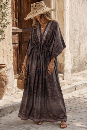 A woman wearing a bohemian-style maxi kaftan dress with a dark brown and purple floral pattern, a matching straw hat, and carrying a woven basket bag, standing on a stone street.