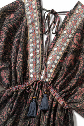 Close-up of a bohemian-style printed dress featuring a dark paisley pattern in black and dusty pink, decorated with rows of silver circular mirror work along the deep V-neckline and tie closure.