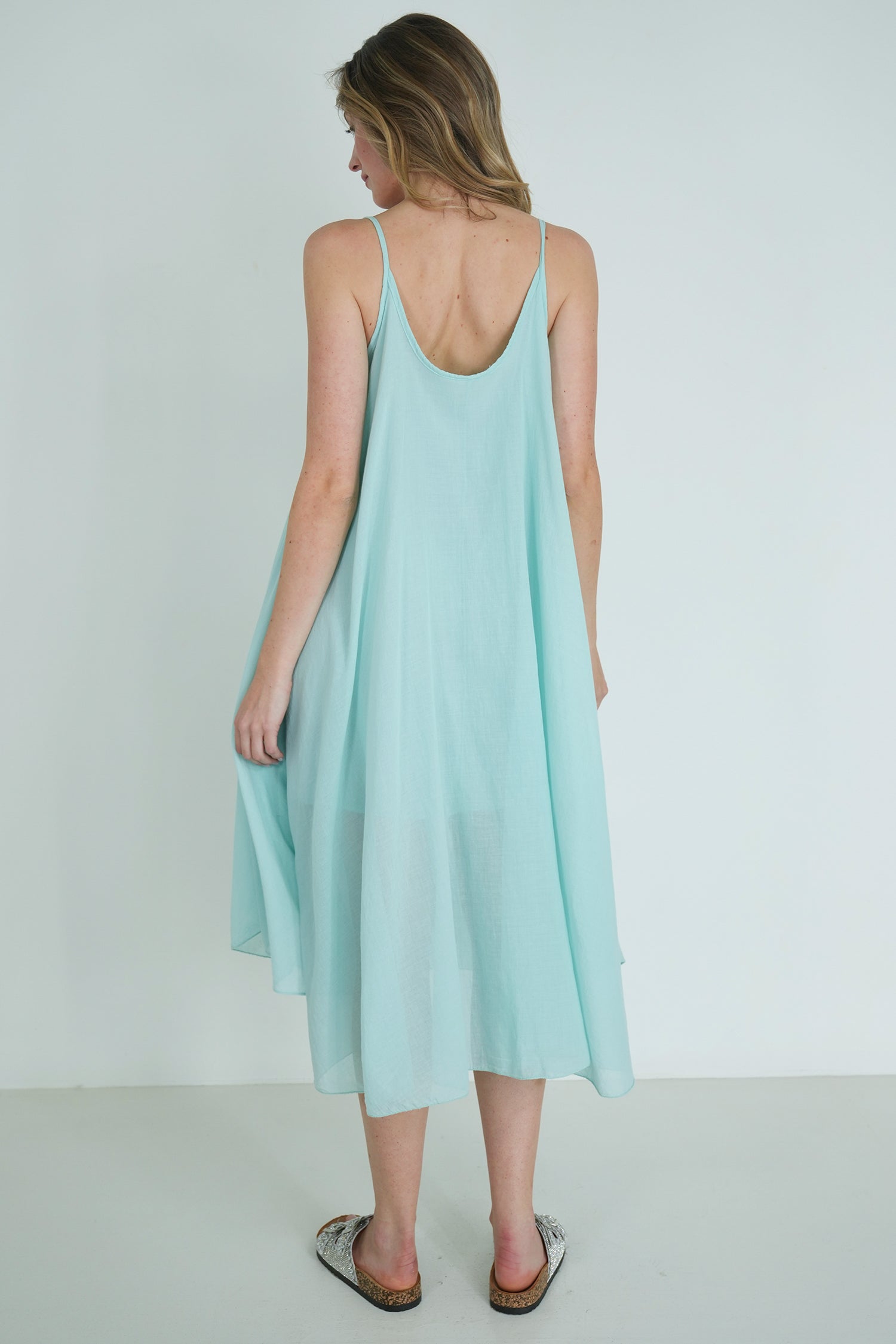 Back view of model wearing a light turquoise sleeveless flowy midi dress with scoop back, styled with silver sandals indoors.