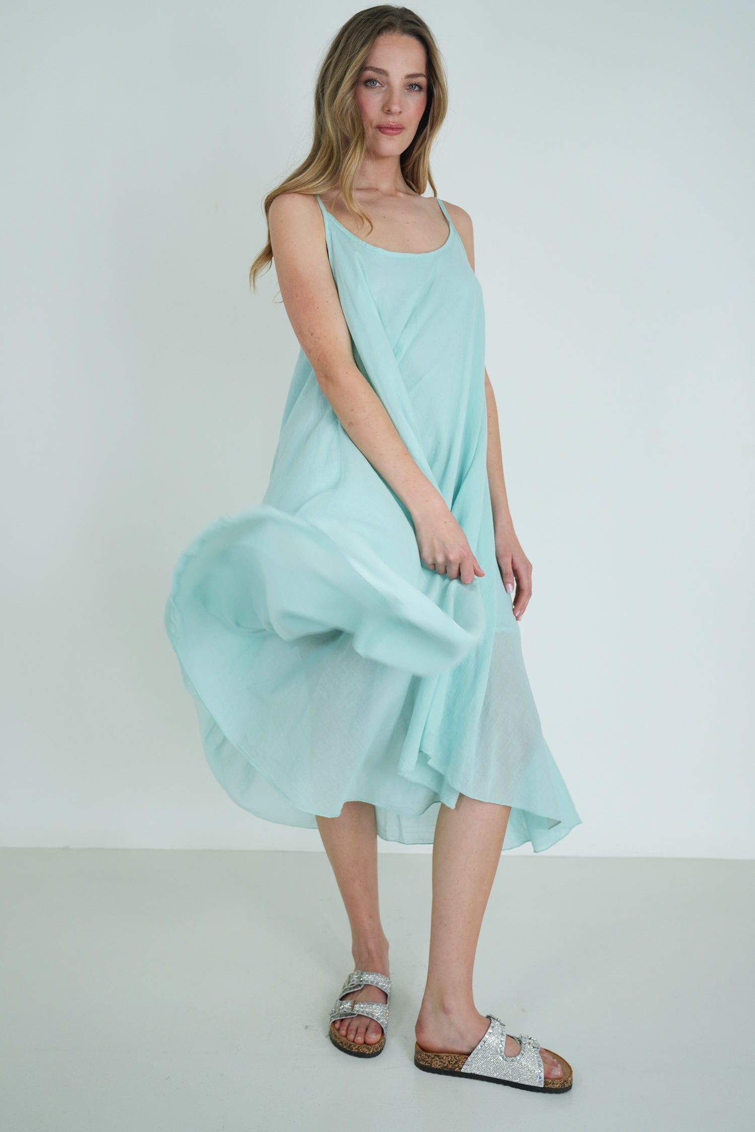 Model wearing a flowing light turquoise sleeveless midi dress with layered hem, styled with silver sandals, posing gracefully indoors.