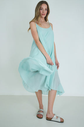 Model wearing a flowing light turquoise sleeveless midi dress with layered hem, styled with silver sandals, posing gracefully indoors.