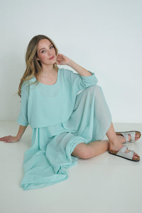 Model sitting gracefully wearing a light turquoise knit top and flowy chiffon skirt, paired with silver sandals against a white backdrop.