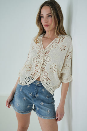 Woman wearing a white lace top and denim shorts against a plain background