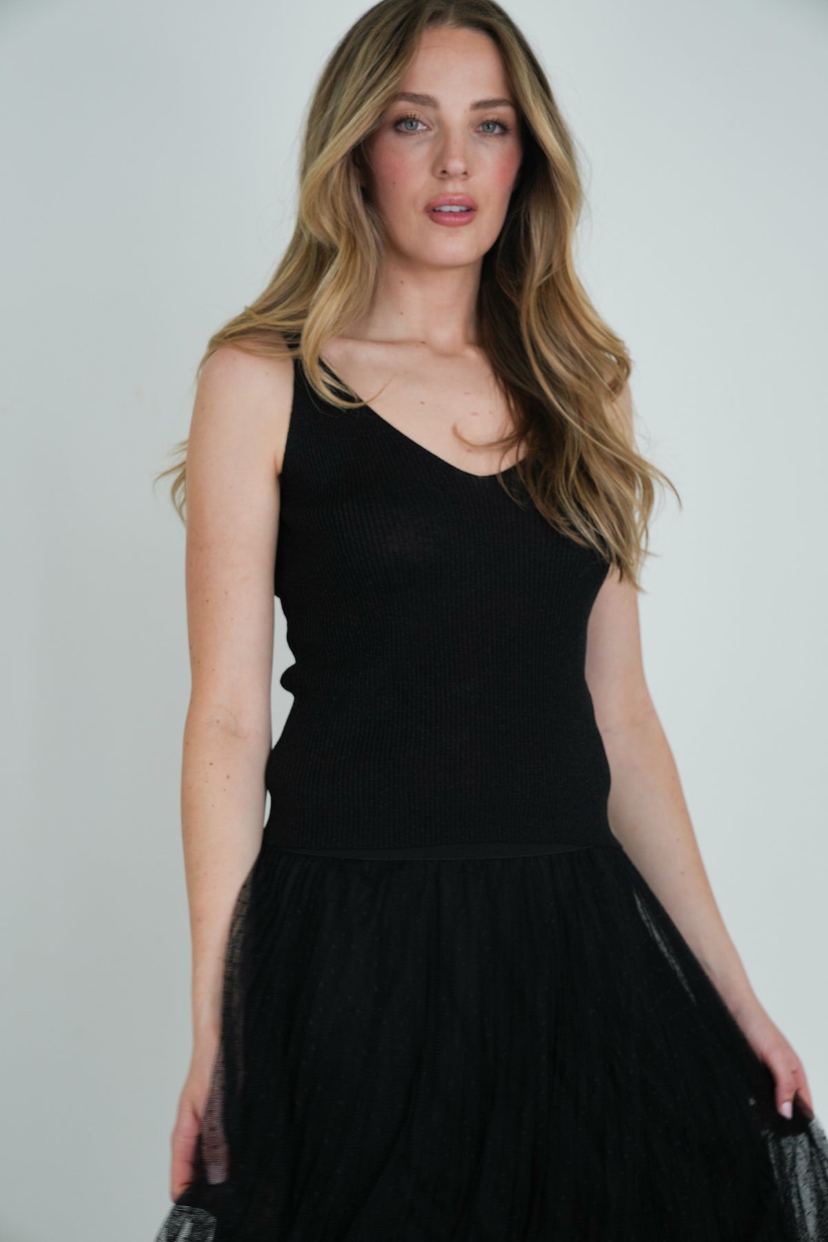 Model wearing a sleeveless black ribbed V-neck top paired with a black tulle skirt, posing against a plain white background."
