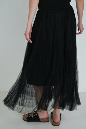 Back view of black pleated tulle midi skirt with sheer layers, showing flowy fabric detail and black sandals with gold buckles.