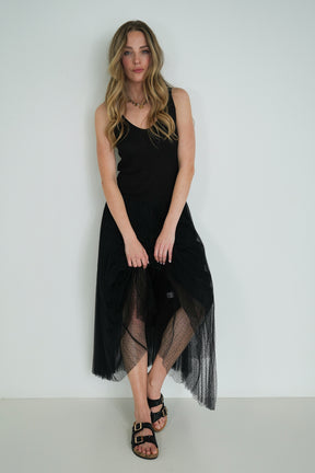 Model wearing a sleeveless black pleated midi dress with sheer tulle overlay, holding the skirt and standing against a white wall.
