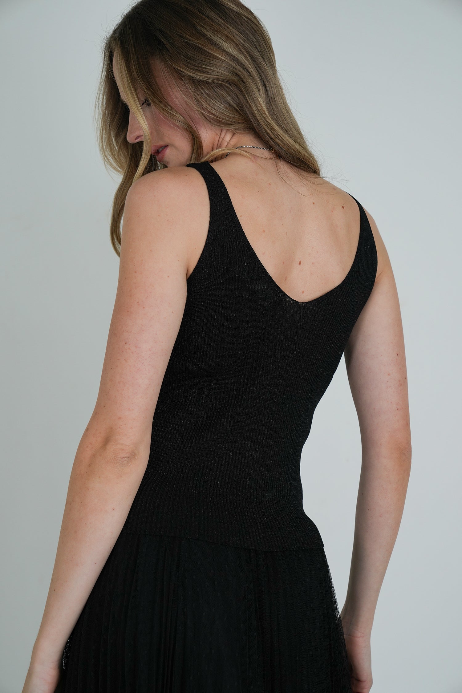 Side-back view of model wearing a sleeveless black ribbed V-neck top and black pleated tulle skirt, showing fitted silhouette.