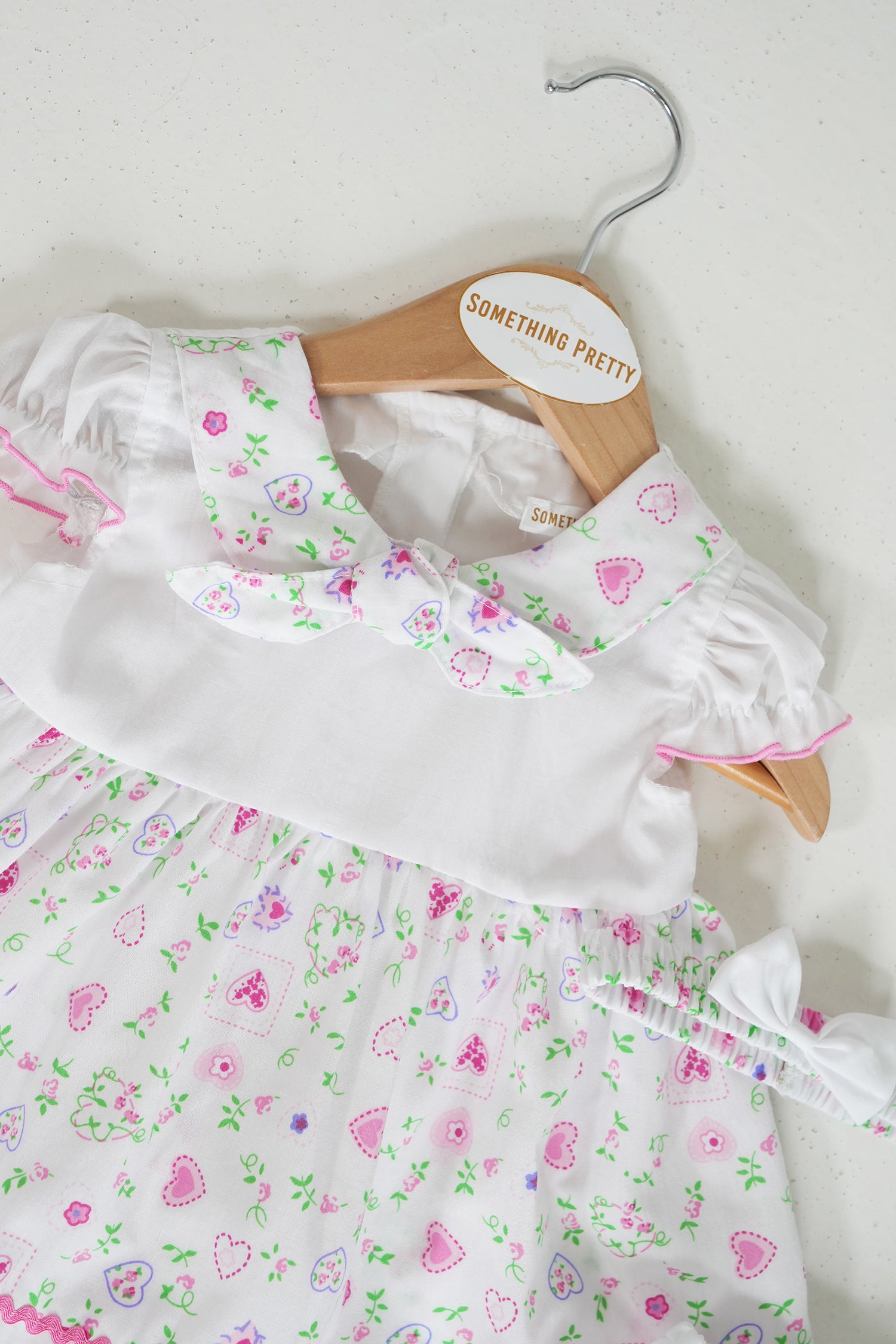 Close-up of White Peggy dress with pink ric-rac trim and matching bow headband, heart and floral print