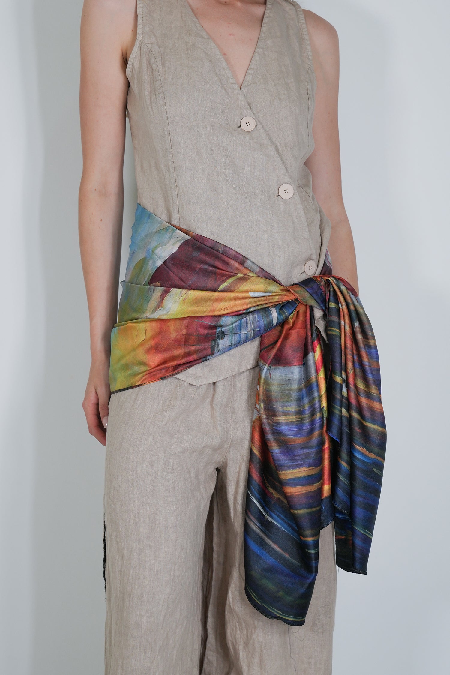 Person wearing a beige sleeveless top and pants with a colourful scarf tied around the waist on a plain background