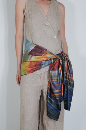 Person wearing a beige sleeveless top and pants with a colourful scarf tied around the waist on a plain background