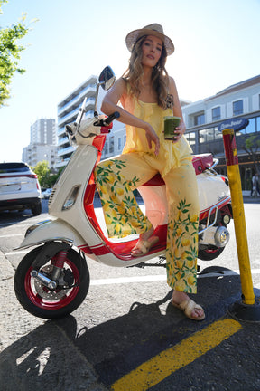 Woman in a yellow dress and hat sitting on a red scooter in an urban setting.