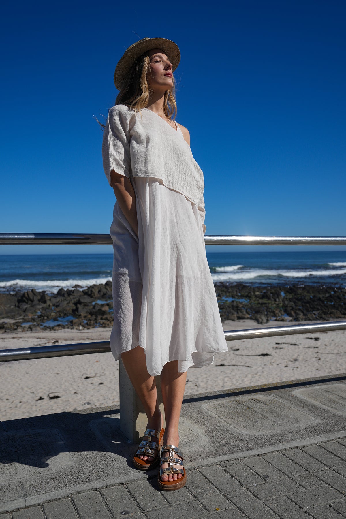 Model wearing off-shoulder white midi dress and sunhat, posing outdoors by the seaside under clear blue sky.