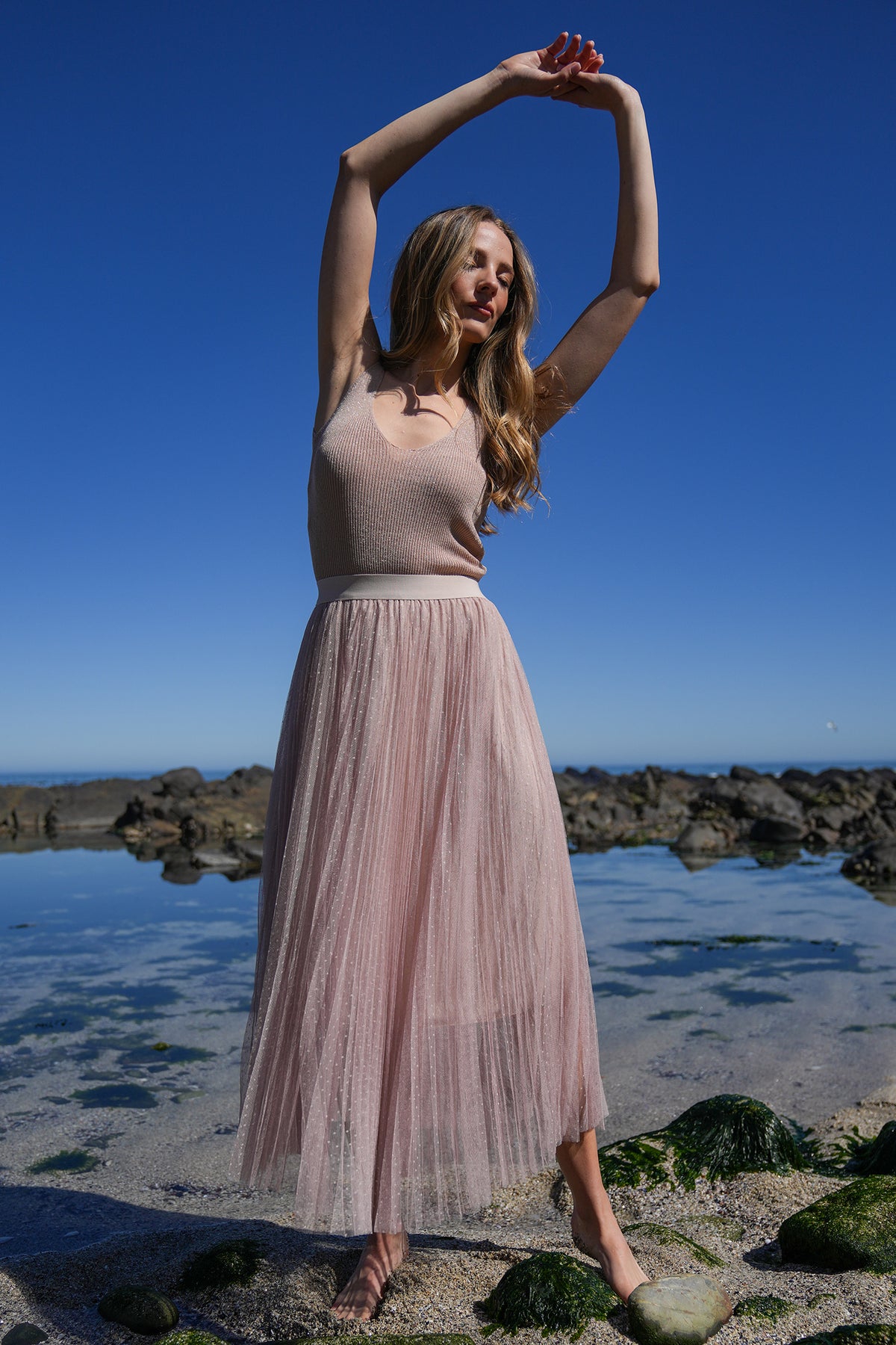 Model wearing a blush pink pleated tulle midi skirt and beige top, posing outdoors near rocks under clear blue sky.