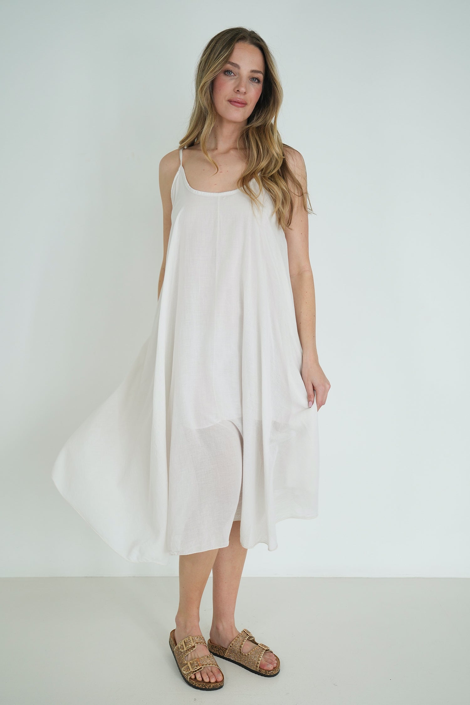 Model wearing a loose white strappy midi dress with flowy fabric, styled with gold sandals, posing against a plain white background.