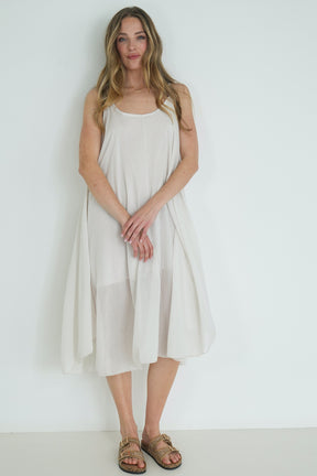 Model wearing a relaxed white sleeveless midi dress with a flowy silhouette and gold sandals, posing against a plain white wall.
