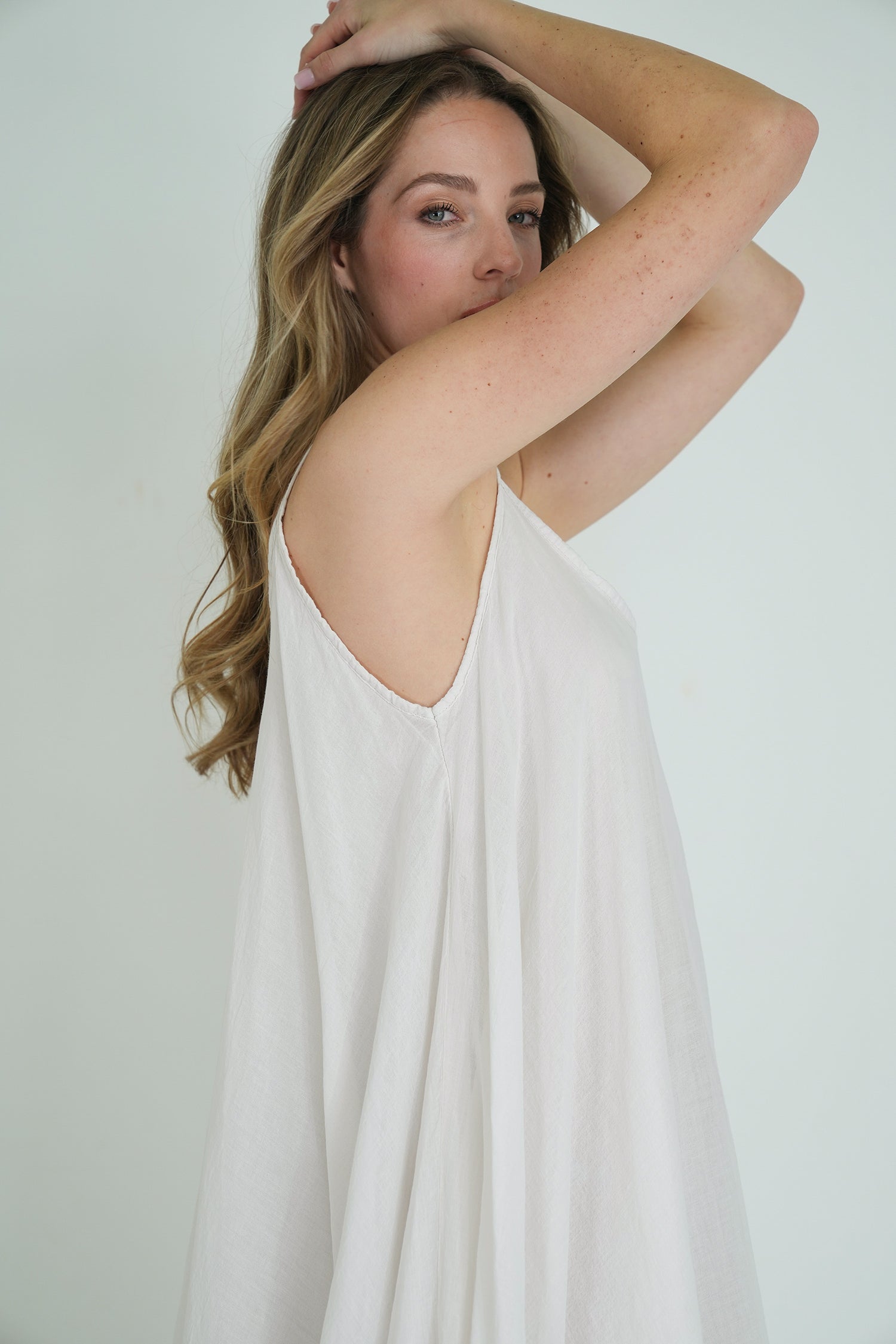 Side view of model wearing a flowy white sleeveless dress, arms raised, showing soft fabric drape and natural elegance.
