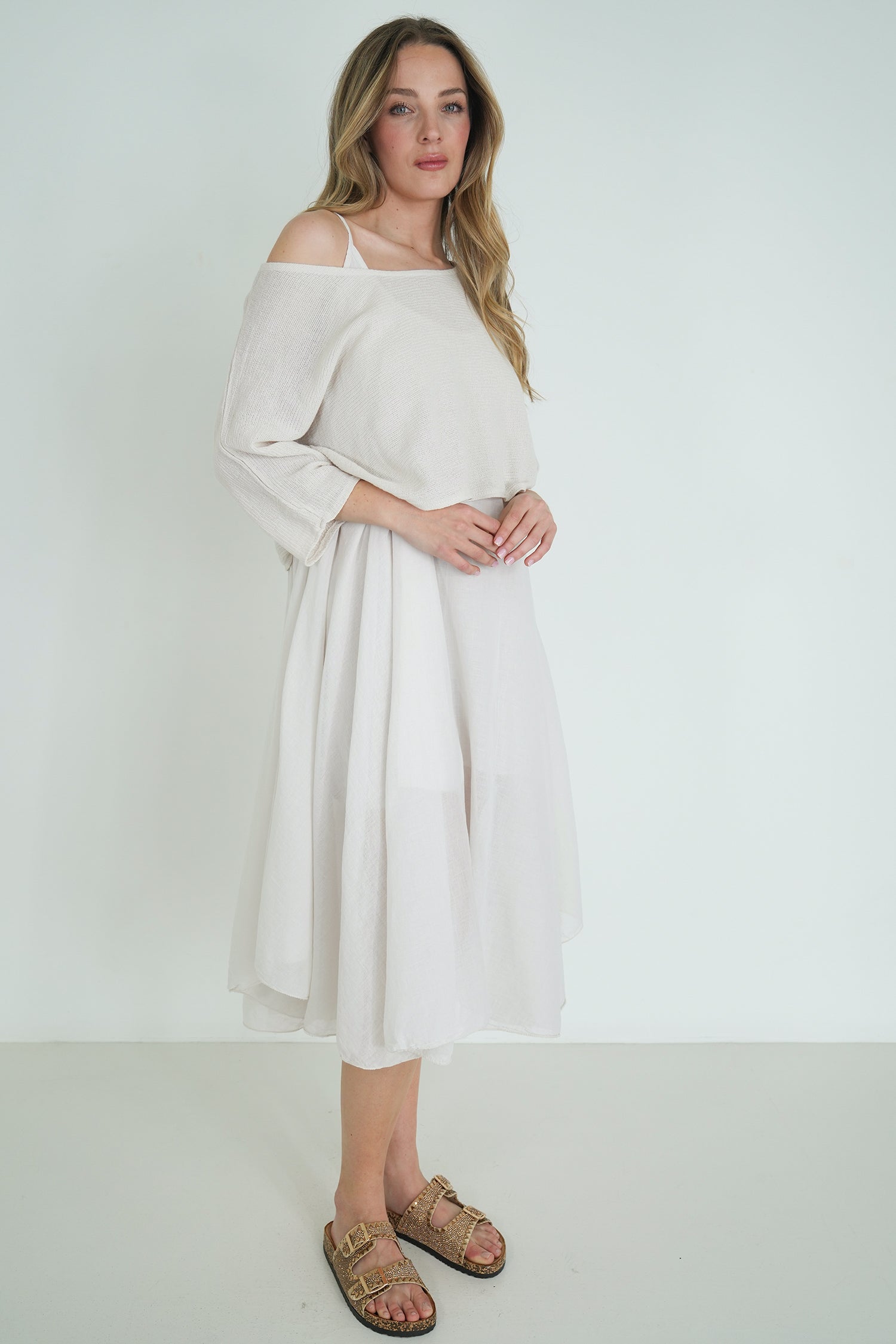 Model wearing off-shoulder cream knit top and flowy white midi skirt, styled with gold sandals, posing against a white background.