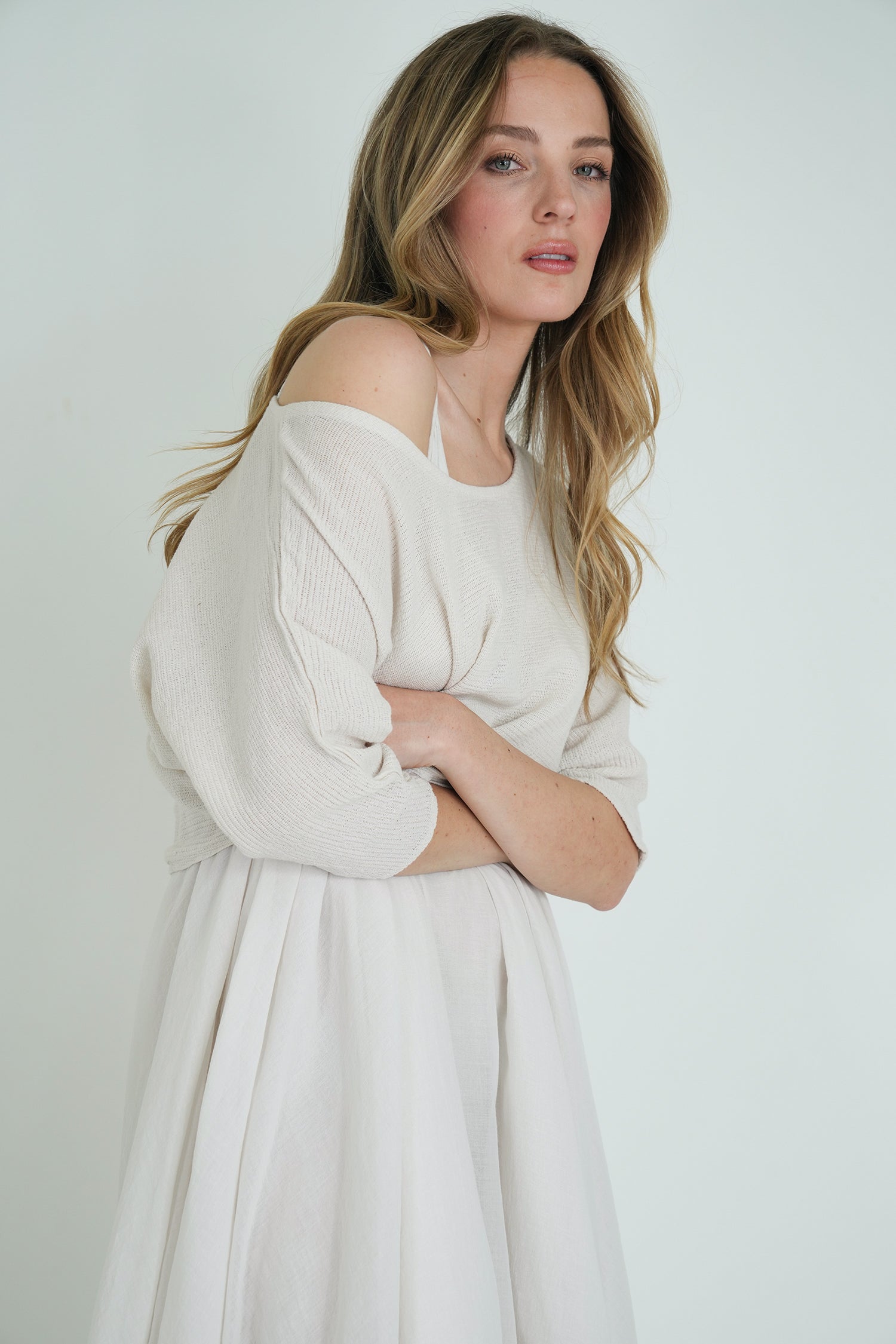 Model wearing an off-shoulder cream knit top paired with a flowing white skirt, posing with arms crossed against a white background.