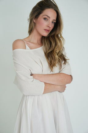Model wearing off-shoulder cream knit top and white skirt, arms crossed, with loose wavy hair against a clean white background.