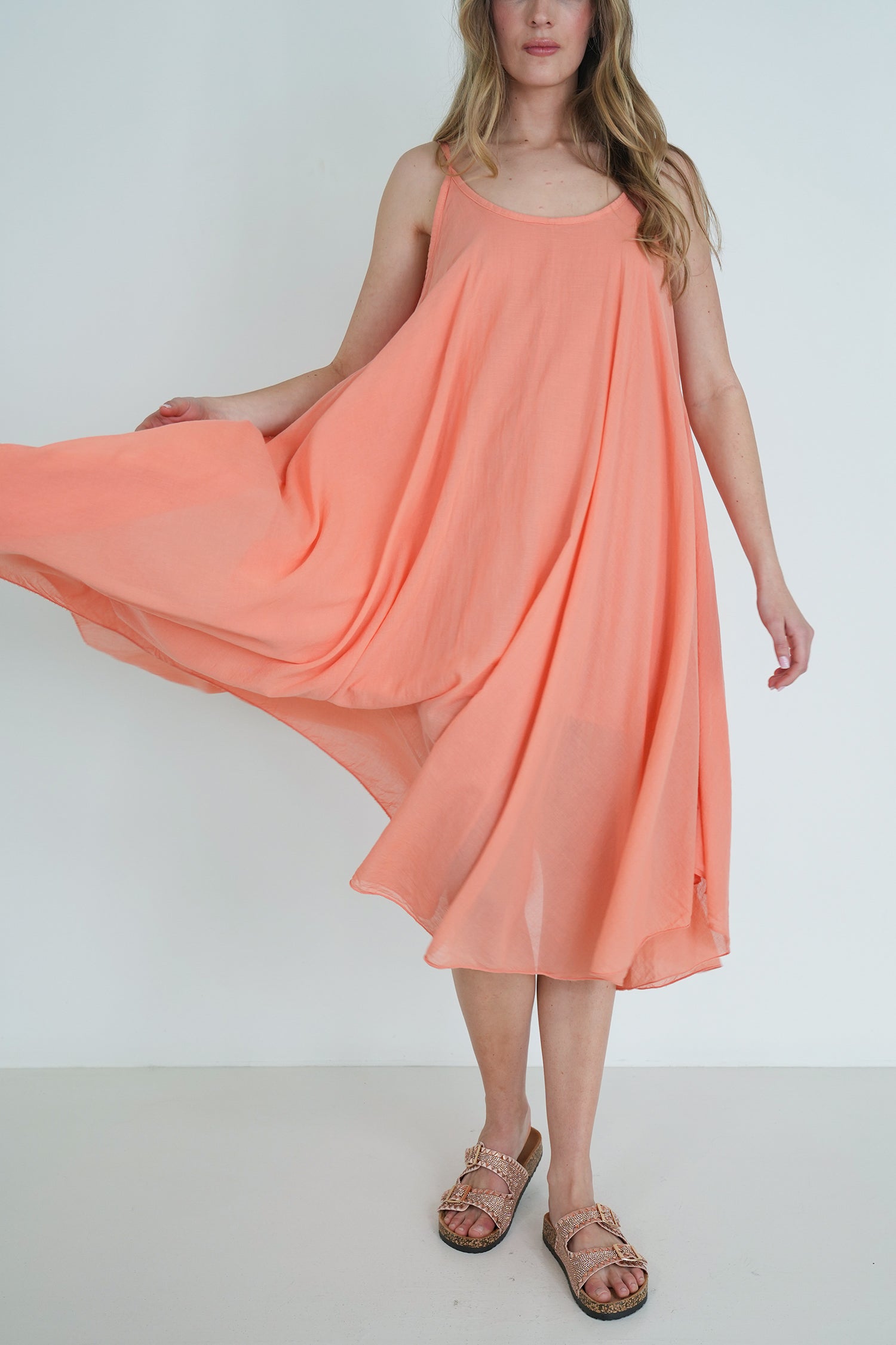 Model wearing a coral sleeveless flowy midi dress with lightweight fabric, holding the skirt to show movement, styled with sandals.