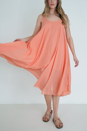 Model wearing a coral sleeveless flowy midi dress with lightweight fabric, holding the skirt to show movement, styled with sandals.