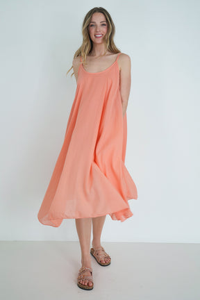 Model wearing a coral sleeveless flowy midi dress with a relaxed silhouette, styled with embellished sandals, smiling indoors.