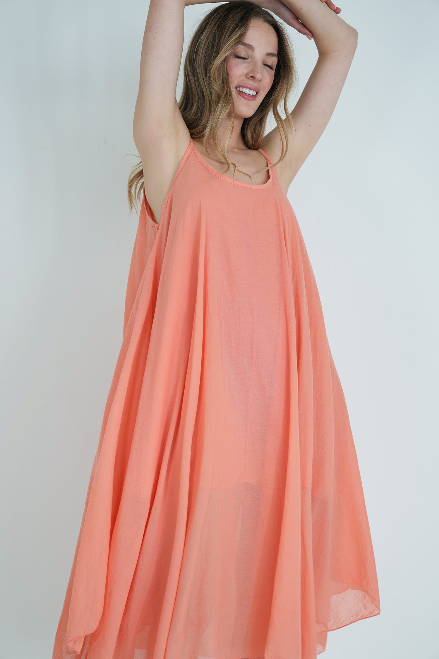 Model wearing a sleeveless coral flowy midi dress with soft pleats, arms raised and smiling, posing against a plain white background.