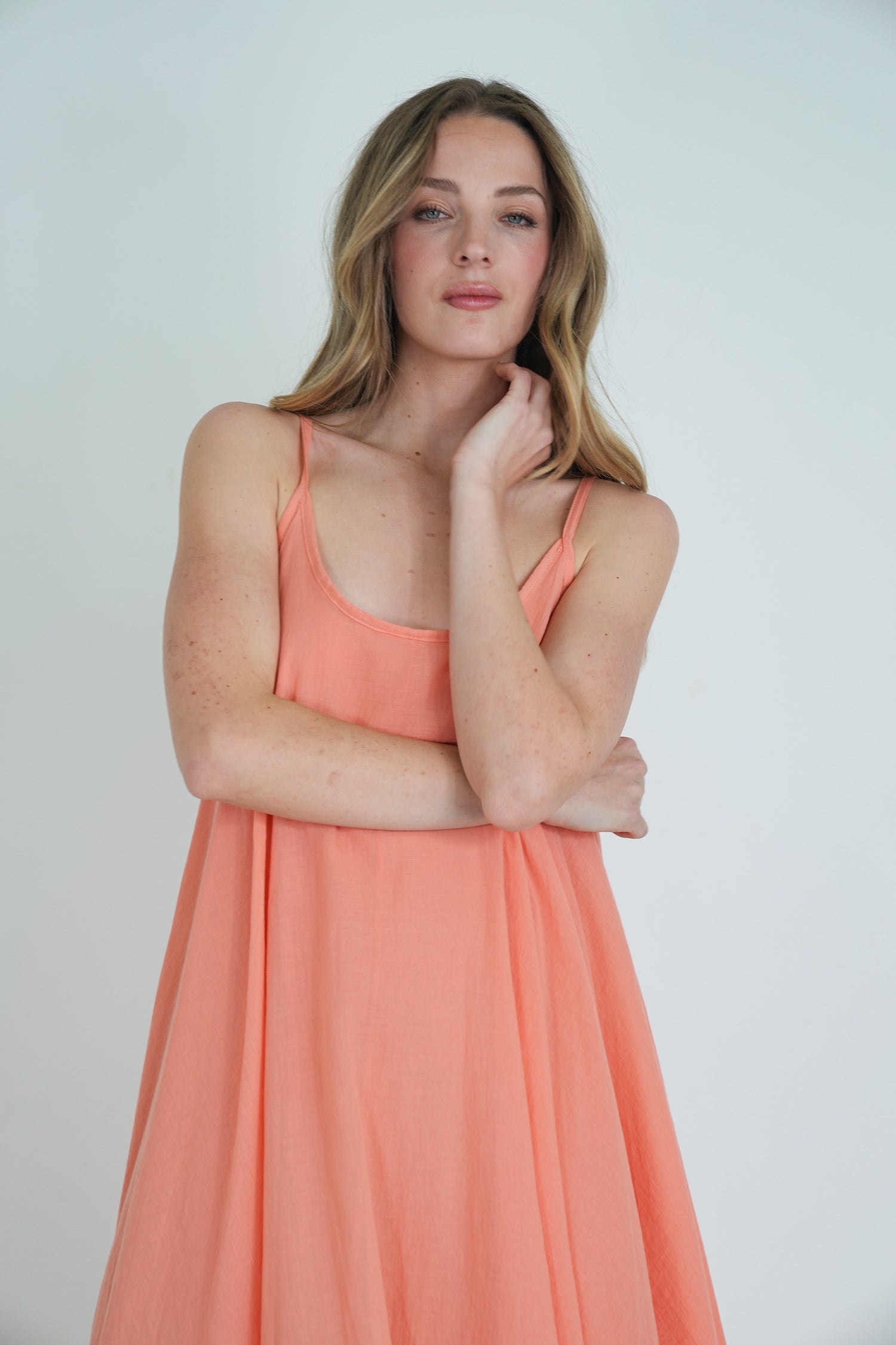 Model wearing a coral sleeveless flowy dress with thin straps, posing softly with one hand on her face against a plain white background."
