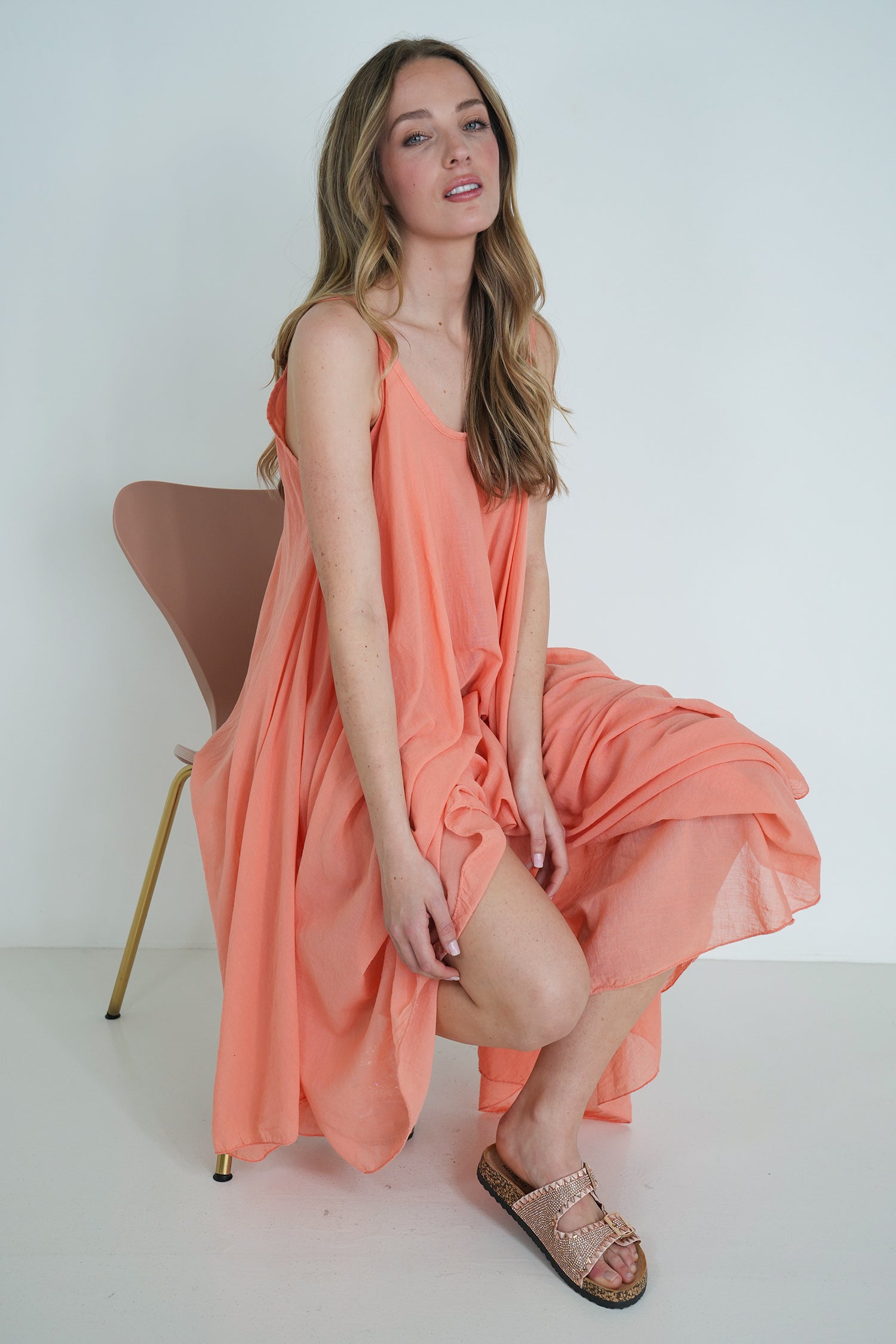 Model wearing coral long-sleeve flowy midi dress with gathered waist, posing with hand on face against white background.