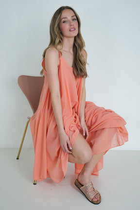 Model wearing coral long-sleeve flowy midi dress with gathered waist, posing with hand on face against white background.