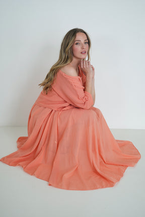Model seated gracefully wearing a coral flowy midi dress with loose sleeves, posing softly against a plain white background.