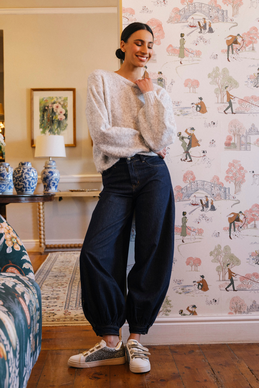A woman stands in a decorated room wearing a fuzzy light gray sweater, dark blue baggy jeans, and white sneakers with gray patterned uppers. The background features a pink and white illustrated wallpaper.