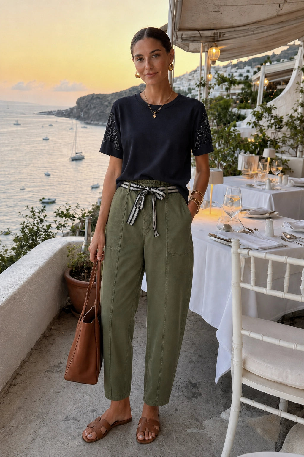 A woman stands on an outdoor terrace overlooking the sea at sunset, wearing a navy short-sleeve top with lace sleeves, olive green drawstring trousers, and brown sandals, while carrying a brown shoulder bag.