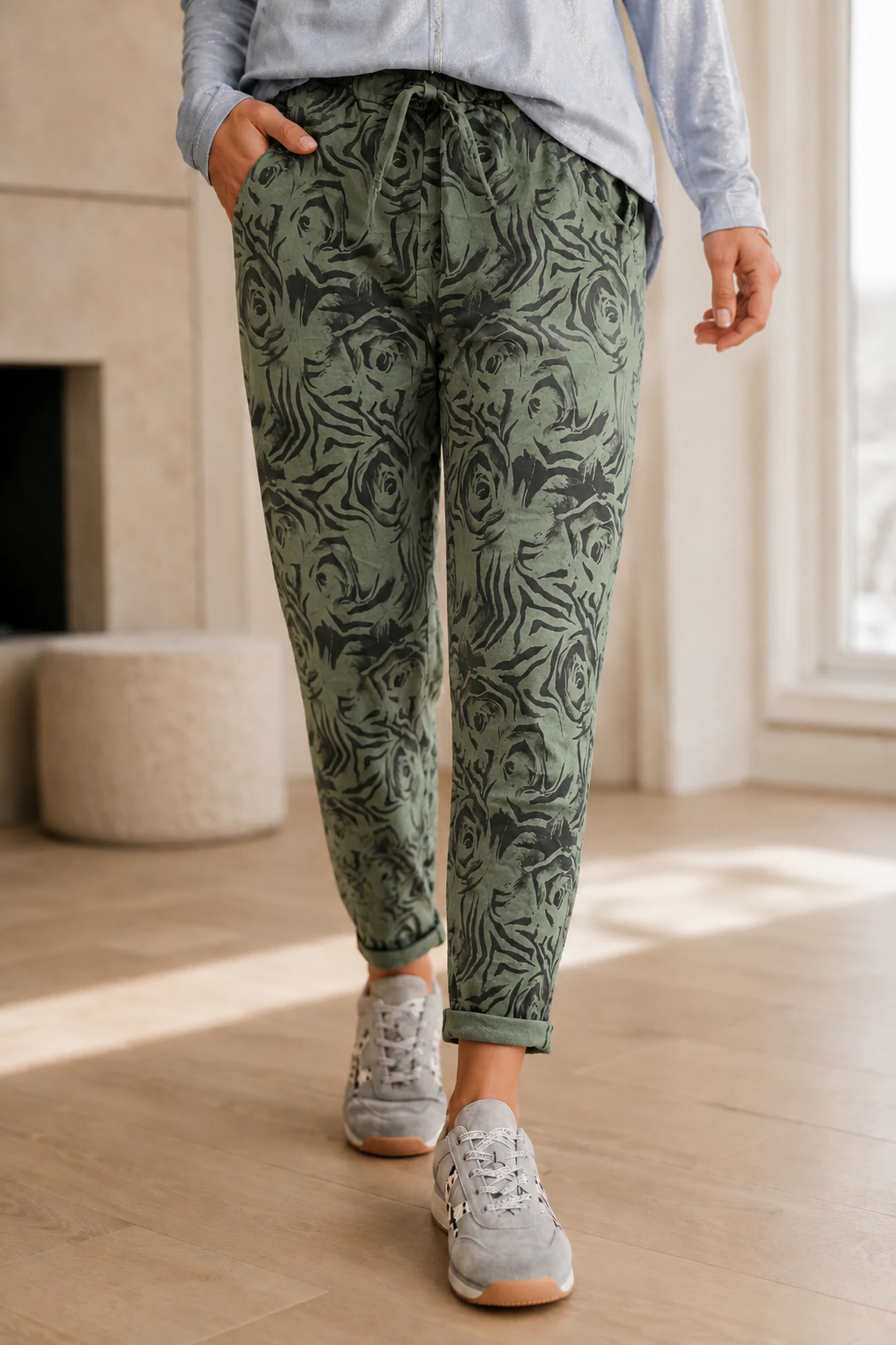 A person wearing sage green drawstring jogger pants with a dark abstract swirl pattern, styled with a light blue long-sleeve top and gray sneakers in a bright indoor room.