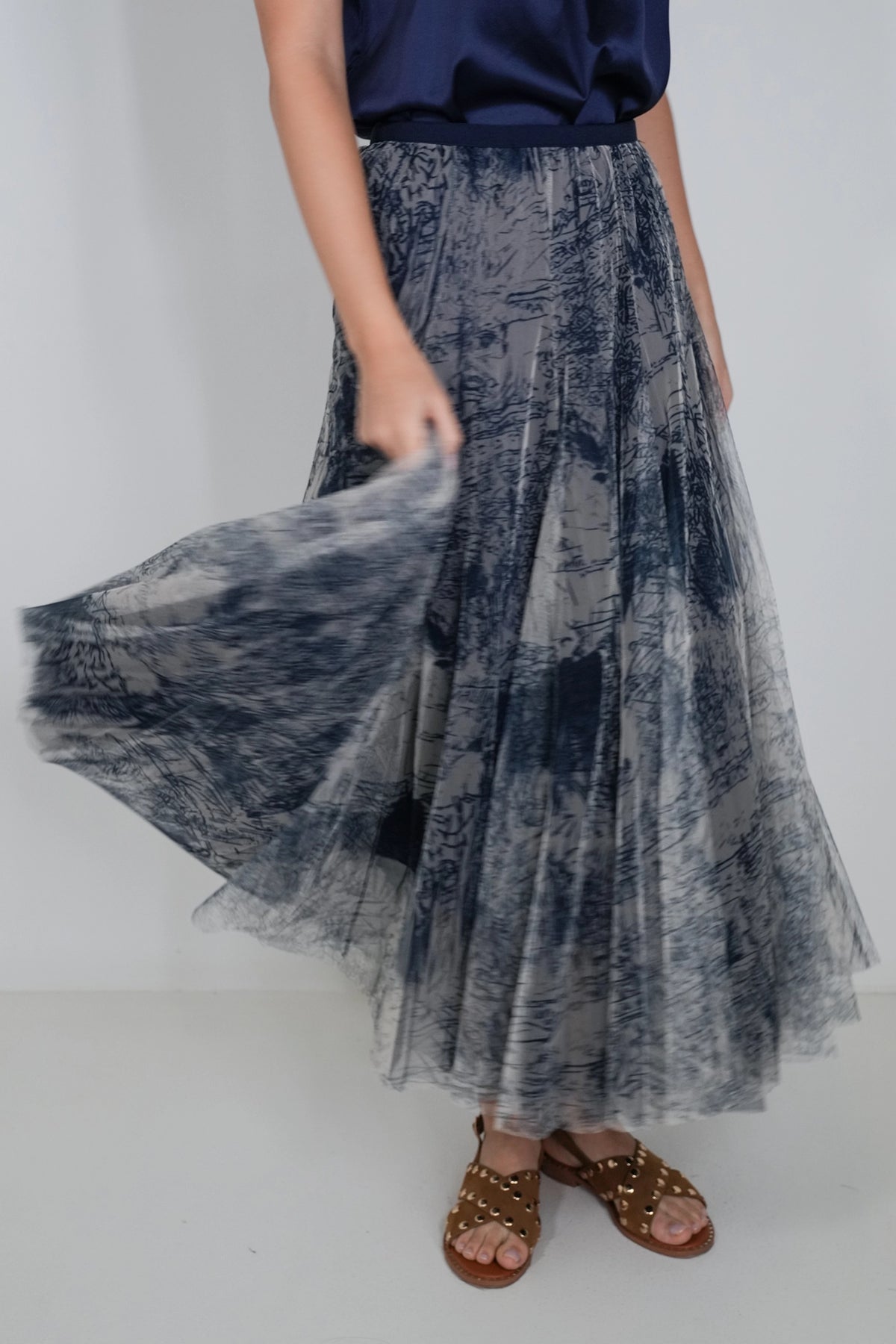 A model wearing a high-waisted, pleated tulle maxi skirt with a blue and gray abstract brushstroke pattern, paired with a navy top and brown studded sandals.