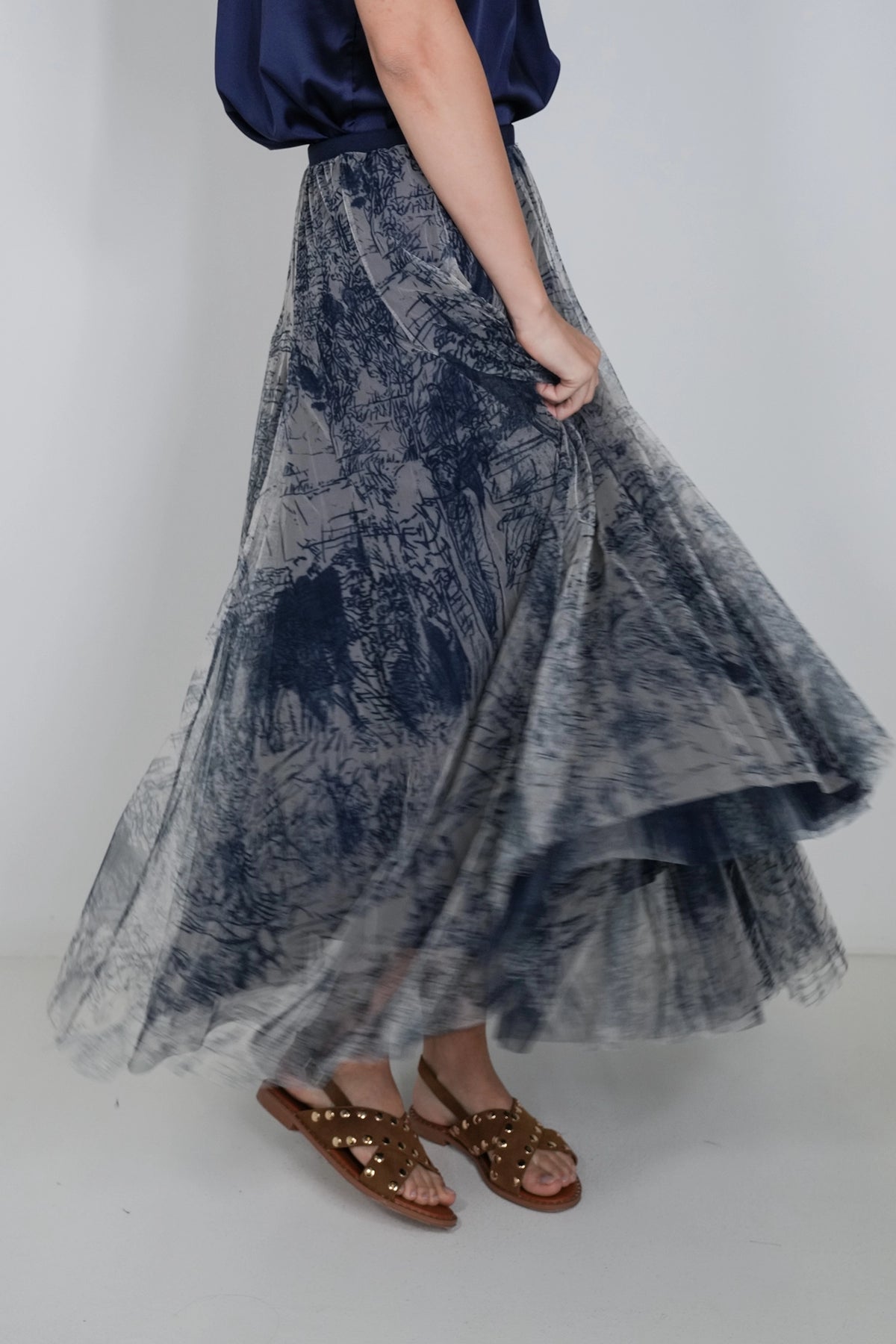 A person wearing a flowing midi skirt with a blue and gray abstract print, paired with brown studded sandals and a navy top, standing against a light gray studio background.